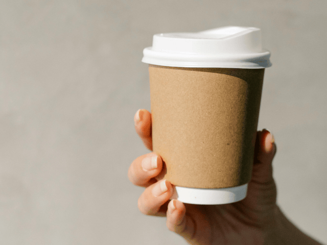 Hand holding a brown paper coffee cup with a white lid against a neutral background